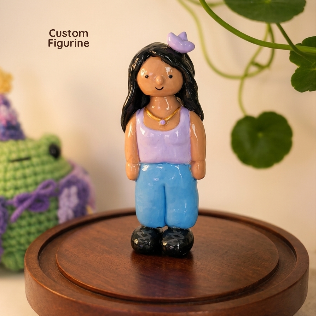 A custom hand-painted clay figurine of a girl with long black hair, a purple top, blue pants, and black shoes, standing on a round wooden display base. The background is softly blurred with a green plant and a crocheted toy.